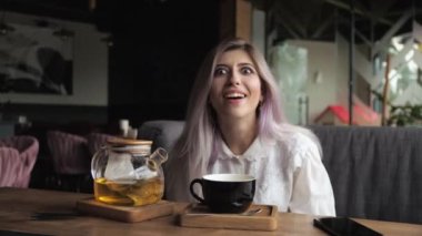 Genç sarışın iş kadını bir restoranda oturur ve pencerenin kenarında çay içer. Şaşırmış, gülümsüyor, dikkatsizce gülüyor, ağzı açık.