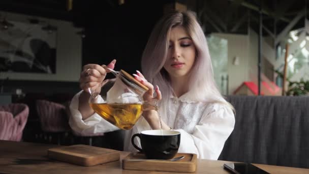 若いブロンドのビジネス女性は透明なガラスのティーポットからカップにお茶を注ぎます レストランでは窓のそばで — ストック動画