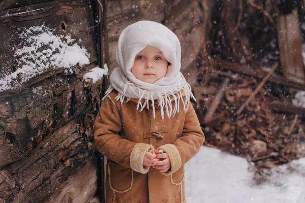 Mała urocza dziewczynka w rosyjskich strojach ludowych stoi na śniegu w pobliżu drewnianego budynku. Dziewczyna w białym szaliku w tle dewastacja, drewno — Zdjęcie stockowe