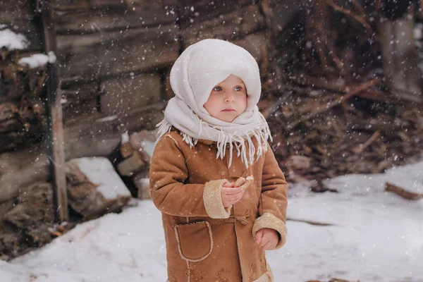 Ein kleines charmantes Mädchen in russischer Tracht steht im Schnee und isst einen kleinen Bagel. Stockfoto