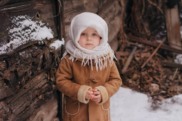 Ein kleines charmantes Mädchen in russischer Tracht steht in der Nähe eines Holzgebäudes im Schnee. Ein Mädchen in einem weißen Schal im Hintergrund Verwüstung, Metall lizenzfreie Stockbilder