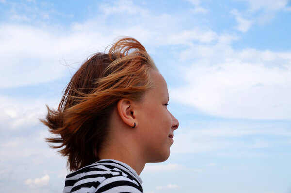 Profile of a smiling red-haired teenage girl against a cloudy sky.