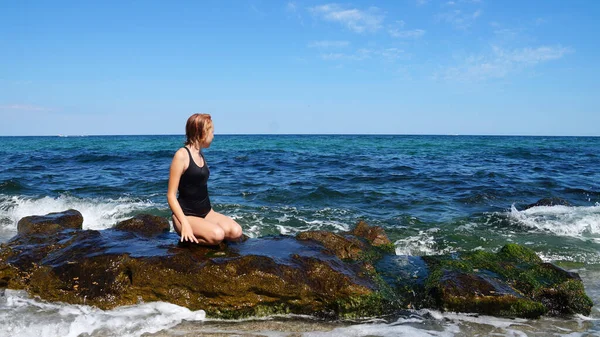 海の地平線に対してビーチに大きな岩の上に座って黒の水着の少女 — ストック写真