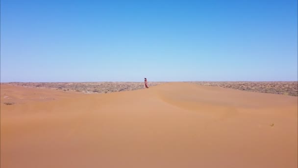 Женщина в красном платье идет по пустыне среди песчаных дюн — стоковое видео