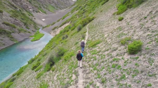 Deux randonneurs, touristes, le voyageur avec des sacs à dos marchent le long de la route de montagne le long du lac turquoise au printemps. — Video