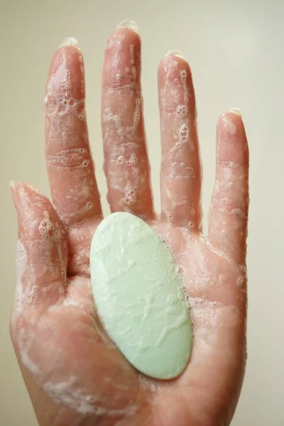Wet female hand in foam hold green bar of soap on a beige background. Side view, closeup.