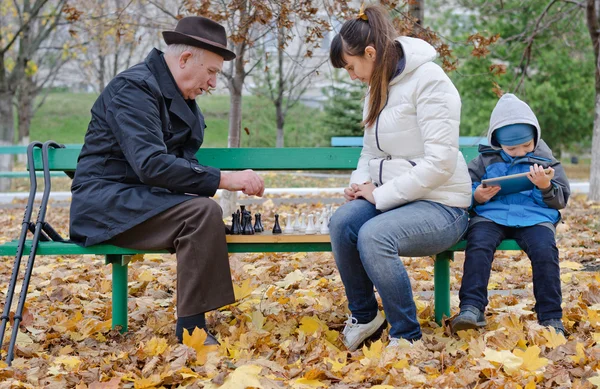 Lindo joven sentado en un banco del parque sosteniendo una tableta mientras su madre y su abuelo juegan al ajedrez — Foto de Stock