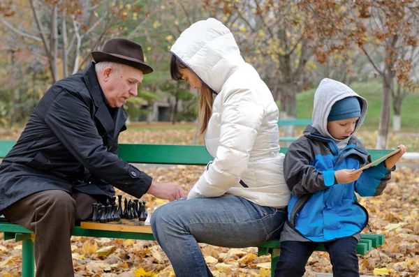 Lindo chico sentado en un banco del parque sosteniendo una tableta mientras su madre y su abuelo juegan al ajedrez —  Fotos de Stock