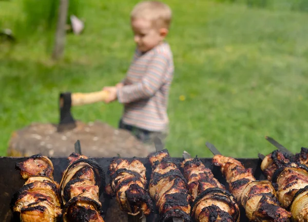 Kebabs grilling on a BBQ as a small boy chops wood — Stock Photo, Image