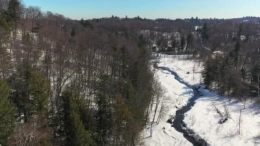Hava aracı videosu yavaşça iniyor ve kır yolunu, dağları ve baharda beyaz karla akan küçük bir dereyi gösteriyor.