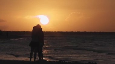 Altın bir günbatımının önünde öpüşen ve kumsalda yürüyen bir çiftin silueti.