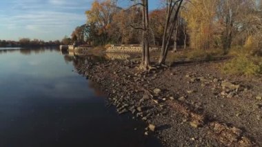 Flying with a drone, mid-air, over the shore of a calm river in the Fall, with naked trees at sunset