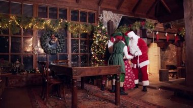 Mutlu elfler Noel Baba 'yı hediyelerle uğurlar. Elfler Noel Baba 'nın sırtına kırmızı bir çuval dolusu hediye koydu. Noel süslemeli ev.