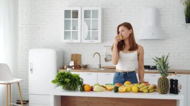 A beautiful sports girl at home in the kitchen eats a fresh apple. Table with different seasonal fruits. Vegetarian healthy food. Diet.