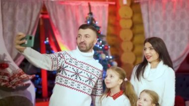 Happy young couple with small children near glowing bright decorated Christmas Tree using smartphone take photography capture moment New Year celebration, family having fun on xmas eve concept
