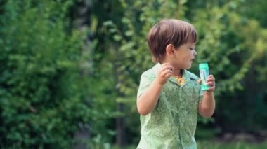 Tişörtlü neşeli bir çocuk zıplıyor, oynuyor ve sabun köpüğü üflüyor. Parkta mutlu bir çocukluk geçirdiniz..