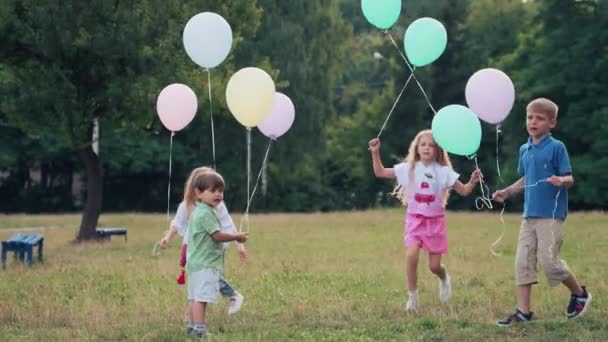 男孩和女孩在公园里奔跑 手里拿着气球的快乐孩子们在草地上玩耍 自然界中快乐而活跃的生日 五颜六色的衣服和气球 — 图库视频影像