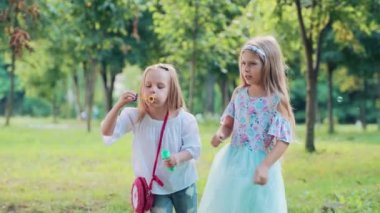 Parkta tatil yapan küçük mutlu kızlar, eğlenin ve sabun köpükleri üfleyin. Güneşli yaz günü, yeşil çimenler ve ağaçlar.