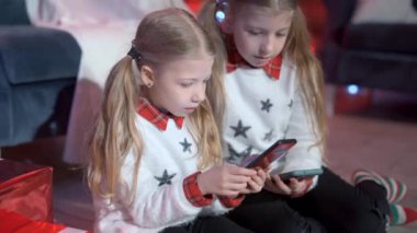 Beyaz Noel kazağı giyen iki tatlı kız kardeş yerde oturup cep telefonlarını ellerinde tutuyorlar. Arka planda Noel süslemeleri.