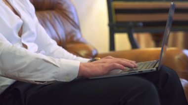 İş adamının elleri dizüstü bilgisayarda klavye kullanıyor. Cep telefonu elinde. Beyaz gömlekli bir adam deri bir kanepeye oturur. Mesai saatleri dışında evden çalış. Yakın plan..