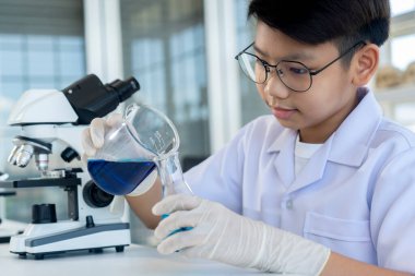 Close up, Portrait Asian boy in glasses pouring blue liquid in beaker into glass bottle in hand with gloves, He smile and looking to blue liquid in chemistry lesson at laboratory