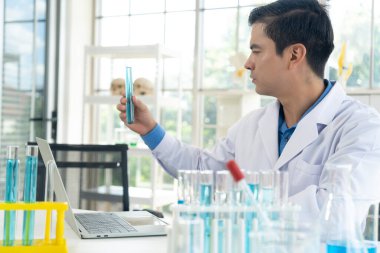 Handsome researcher man in white coat holding and looking on test tube to analyzing blue liquid with laptop computer while working in the laboratory