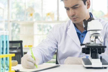Young scientist man in lap coat and gloves working with microscope and write on workpaper during researching chemistry at laboratory