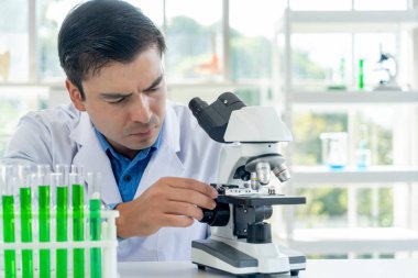 Young male researcher in lab coat looking through microscope with thoughtfully while working in the laboratory, copy space