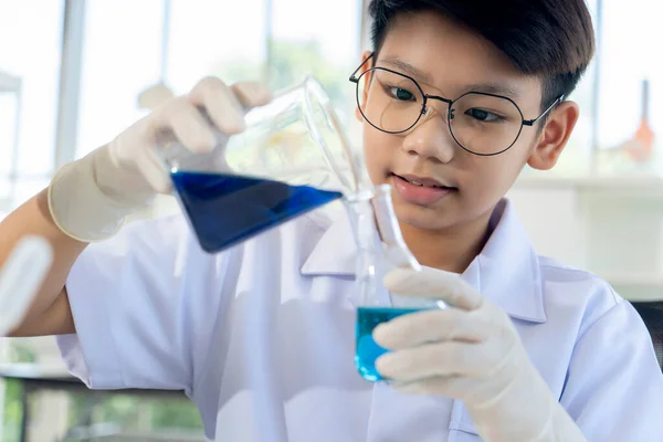 Selective focus Face of Asian boy in glasses pouring blue liquid in beaker into glass bottle in hand with gloves, He smile and looking to blue liquid in chemistry lesson at laboratory