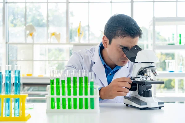 Young male researcher in lab coat looking through microscope with thoughtfully while working in the laboratory, copy space