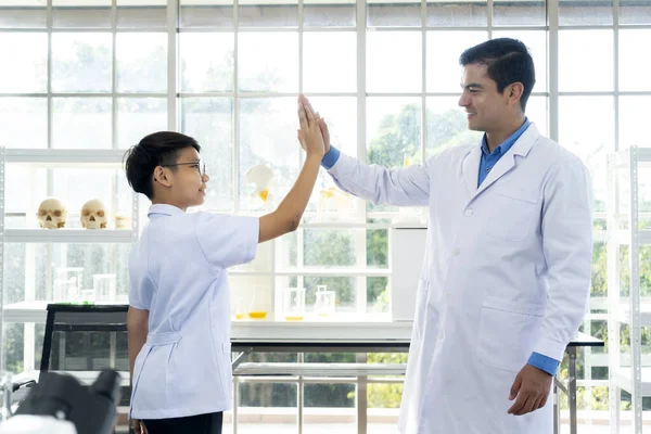 young teacher and asian male student in white coat standing with smile and high five,  They are looking together with feels happy in successful in laboratory at school
