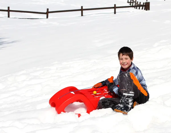 Милий хлопчик біля червоних санчаток в горах на білому снігу — стокове фото