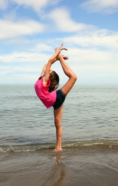 年轻的高加索女孩在海滩和大海的海岸上做有节奏的体操练习 这是一项非常困难的单腿运动 — 图库照片