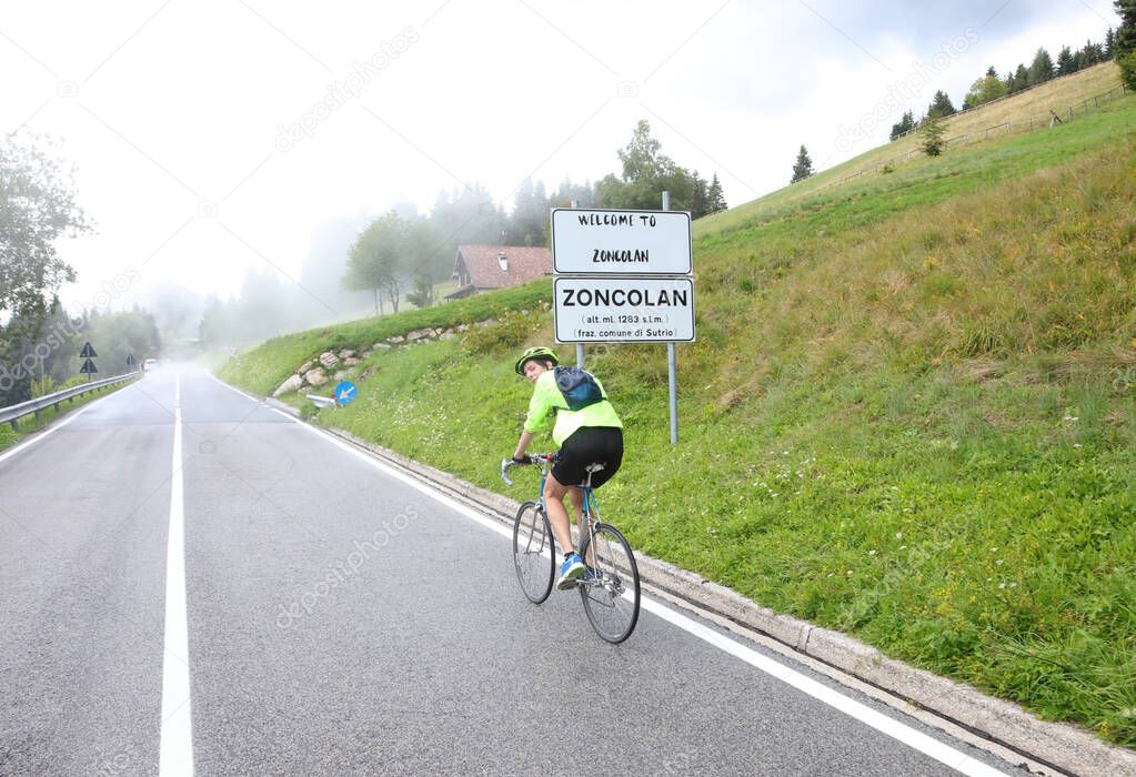 young cyclist with protective helmet pedaling laboriously on the uphill road to Monte Zoncolan in Italy with rain gear during a rainy day