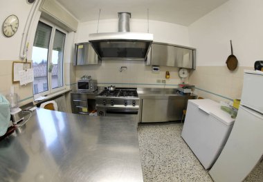 Büyük bir sanayi mutfağının içinde soba ve büyük paslanmaz çelik kaputu ve balık gözü mercekli bir dondurucuyla fotoğraflanmış.