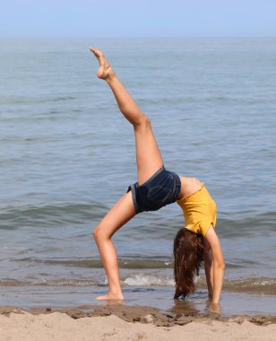 Deniz kenarında jimnastik yaparken sırtı kemerli, bacağı yüksek sarı tişörtlü ince bir kız.
