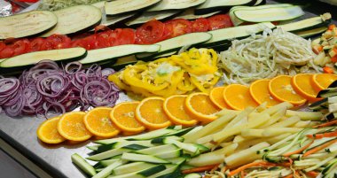 Endüstriyel kantin restoranında yemek hazırlamak için kesilmiş sebzeler ve portakal dilimleriyle dolu çelik bir masa.