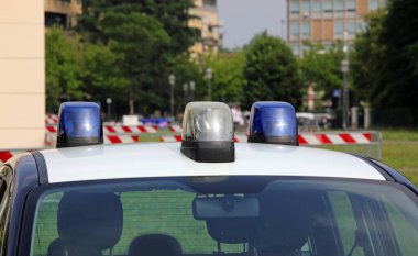 Şehirde mavi ışıklı sirenli bir polis arabası ve arka planda evleri ve binaları var.