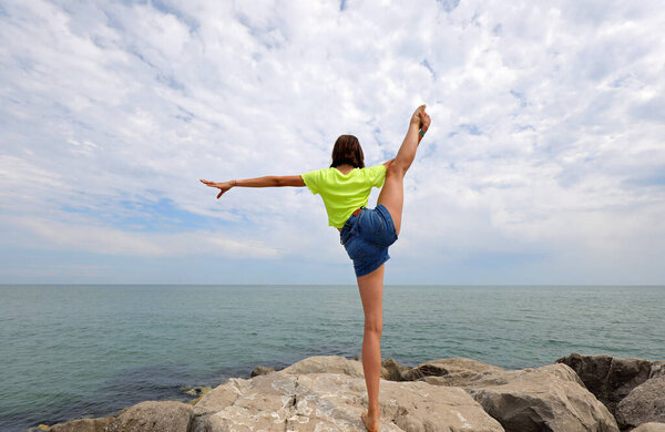 young girl does rhythmic gymnastics exercises on the rocks by the sea with jeans shorts and a fluorescent t-shirt in summer