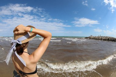 young caucasian girl with hand on straw hat looks at the sea from the shore