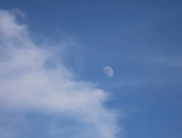 white moon just risen and barely visible during the day in the blue sky and the clouds