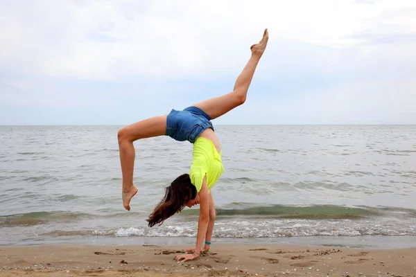 頭を逆さまにして体操をしながら海の岸の細い女の子 — ストック写真