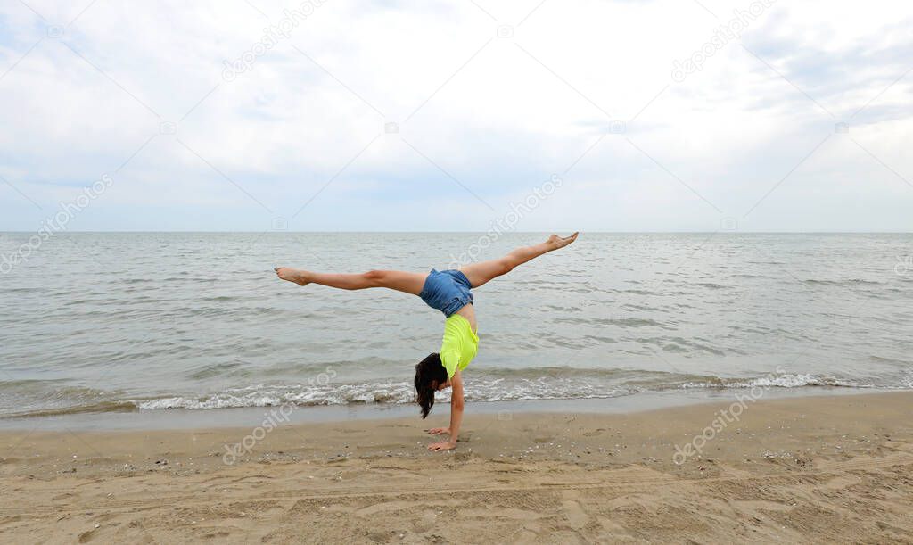 slender teenage girl performs a split with long legs on the seashore in summer