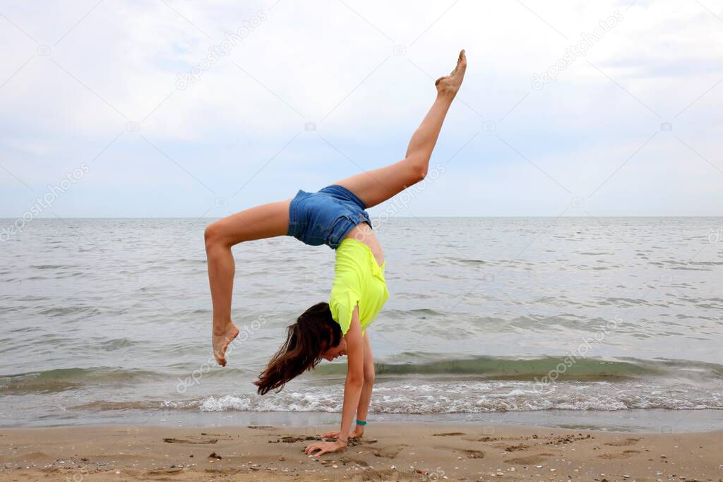 slender girl on the foreshore of the sea while performing gymnastic exercises with her head upside down