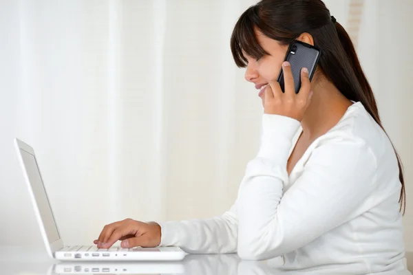 Young woman using her laptop speaking on cellphone — Stock Photo, Image