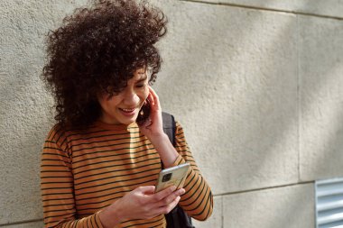 Güler yüzlü siyah bir kız cep telefonuyla dışarıda tuğla bir duvarın önünde dikiliyor.
