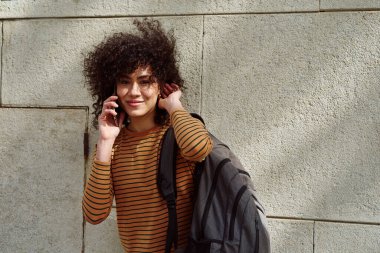 Güzel siyah kız dışarıda tuğla bir duvarın önünde beklerken telefonla konuşuyor.