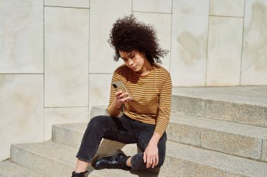 Güneşli bir günde merdivenlerde tek başına oturmuş cep telefonundan bir şeyler okuyan sevimli siyah kız.