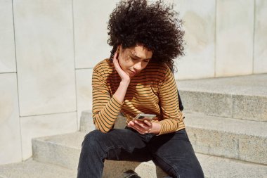 Çekici siyah kız güneşli bir günde tek başına merdivenlerde otururken cep telefonundan bir şeyler okuyor.