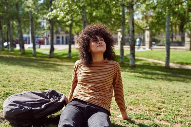 Güneşli bir günde parkta tek başına otururken bakışlarını kaçıran sevimli siyahi kadın.
